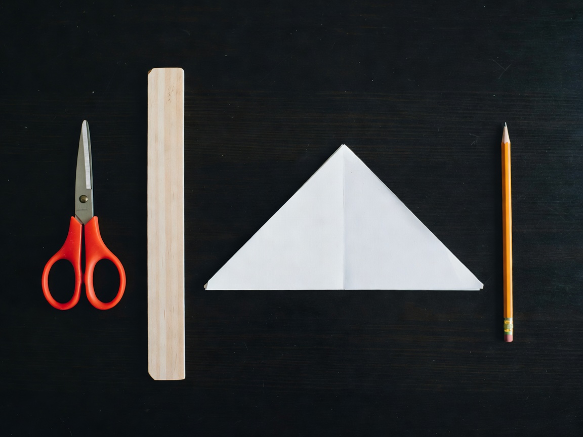 Scissors, a ruler, folded paper and a pencil arranged neatly on a dark beginner craft workspace